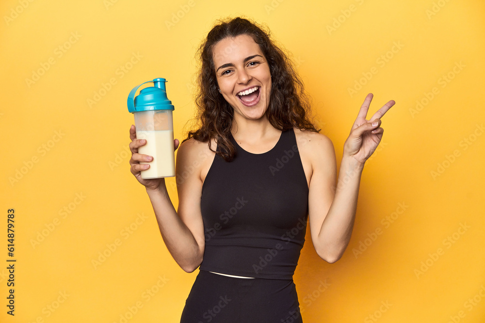 Woman holding a protein shake, in sportswear, joyful and carefree showing a peace symbol with fingers. Woman holding a protein shake, in sportswear, joyful and carefree showing a peace symbol with fingers.