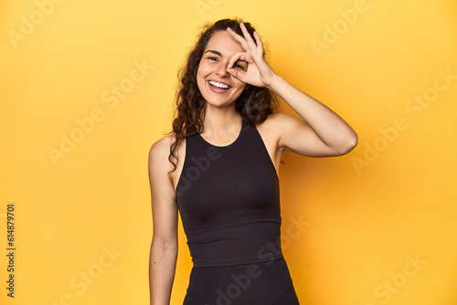 Sporty woman in active wear, yellow backdrop, excited keeping ok gesture on eye.