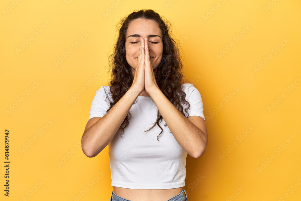 Young Caucasian woman, yellow studio background, holding hands in pray near mouth, feels confident. Young Caucasian woman, yellow studio background, holding hands in pray near mouth, feels confident.