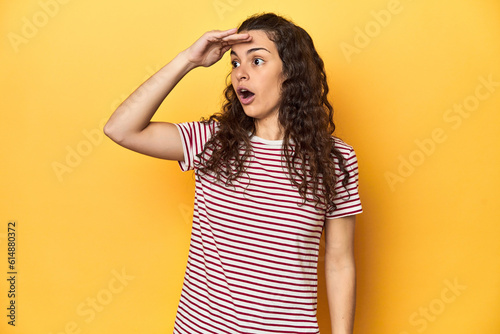 Young Caucasian woman, yellow studio background, looking far away keeping hand on forehead.