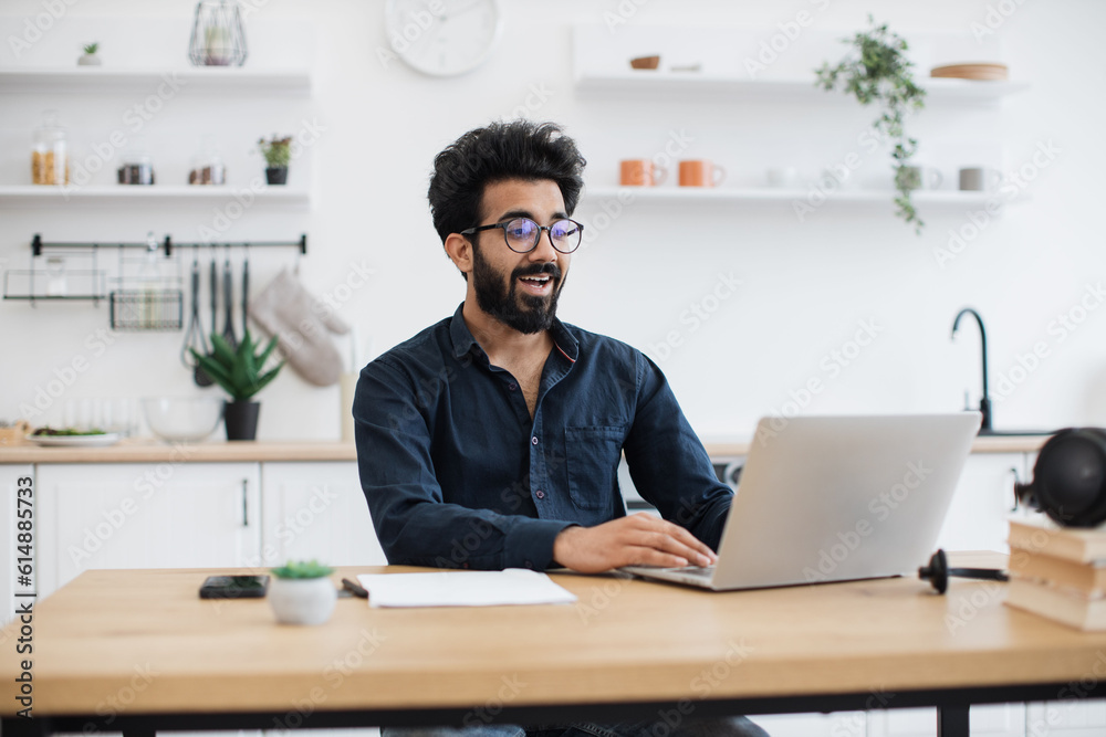 Joyful bearded man in glasses operating modern laptop while sitting at writing desk in spacious dining room. Smiling indian freelancer using flexible schedule while spending working hours at home.