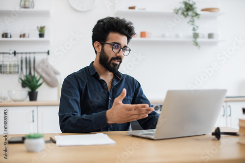 Serious young man in business casual clothes holding online conference with coworkers from home office. Focused indian entrepreneur discussing company issues using portable computer in apartment.