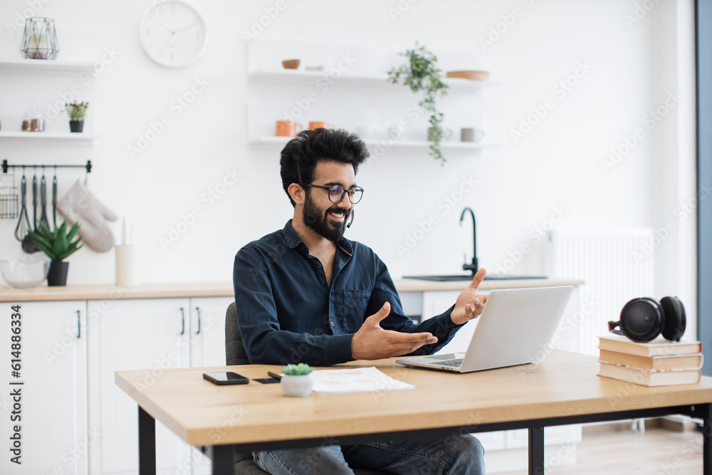 Mindful arabian male in business casual clothes using microphone of headset while holding online briefing via laptop. Efficient remote employee informing colleagues about working arrangement at home. Mindful arabian male in business casual clothes using microphone of headset while holding online briefing via laptop. Efficient remote employee informing colleagues about working arrangement at home.