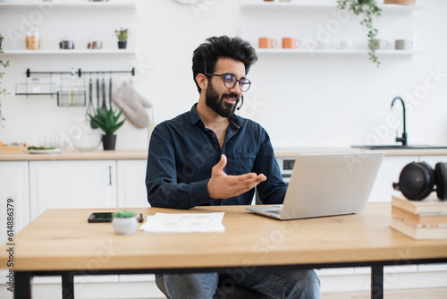 Fotografie Mindful arabian male in business casual clothes using microphone of headset while holding online briefing via laptop