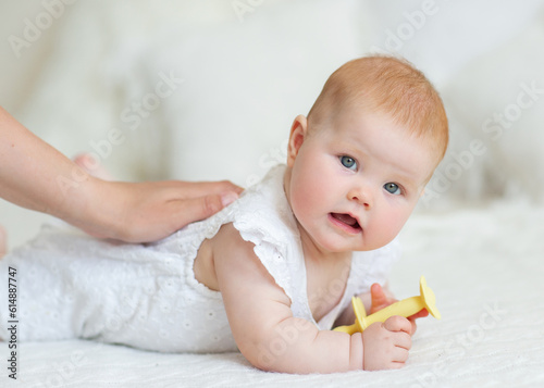 The concept of childcare. Happy little red-haired girl playing on a white blanket in a sunny nursery. The baby lies on his stomach and gnaws teethers. High quality photo