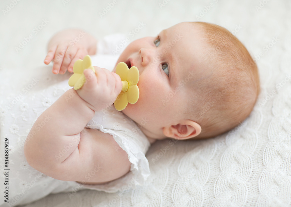 The concept of childcare. Happy little red-haired girl playing on a white blanket in a sunny nursery. The baby lies on his stomach and gnaws teethers. High quality photo