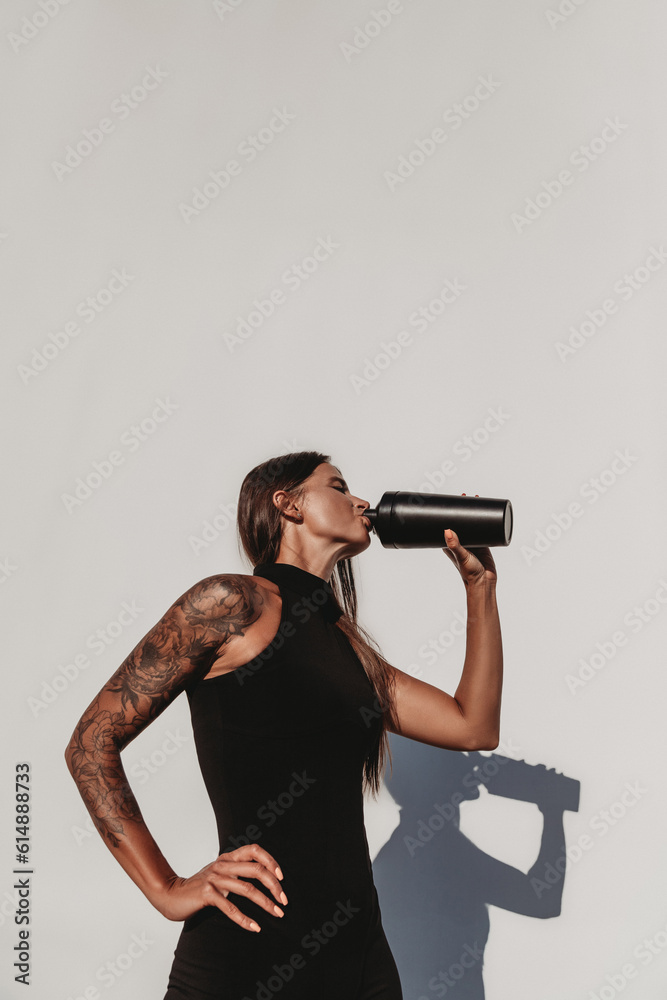 Beautiful young fit tanned tattooed caucasian woman with a protein shaker or water bottle outdoors