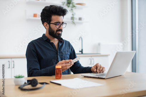 Anxious adult person holding round pills while having online conversation via computer on kitchen background. Aching indian man receiving doctor's consultation via internet connection at home. photo