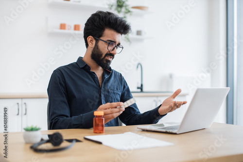 Anxious adult person holding round pills while having online conversation via computer on kitchen background. Aching indian man receiving doctor's consultation via internet connection at home.