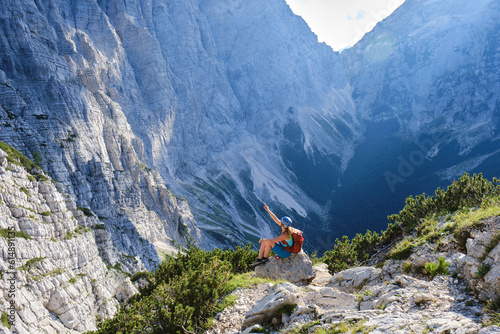 Impressive scale of mountains in Triglav National Park, Slovenia. Woman on via ferrata sits on a rock and points up, towards the top. Summer, adventure, active.