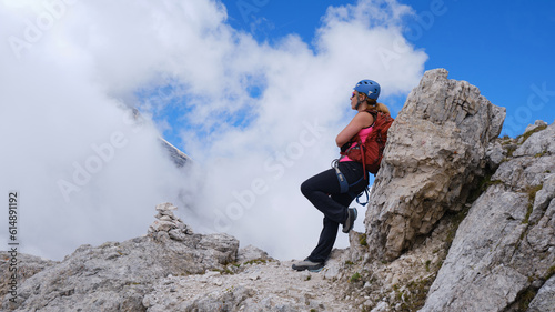Woman rests with her back against a rock on a via ferrata route in Dolomites mountains, Italy, a small rock cairn in front of her and white clouds around. Summer activities, adventure.