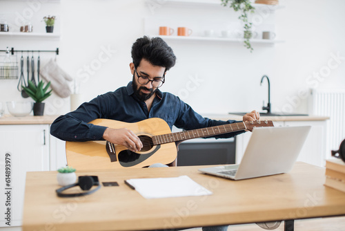 Young male musician in dark shirt tuning acoustic guitar using laptop on writing desk in modern apartment. Focused indian male practising audio exercises on string instrument via online tutorial.