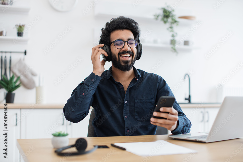 Passionate young man wearing dark shirt and glasses enjoying loud music in headphones while relaxing in dining room. Happy arabian adult using cell phone application for tracks download indoors.