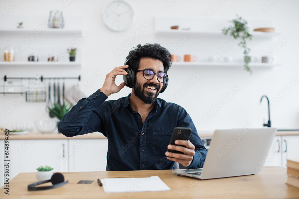 Easygoing arabian person in spectacles showing great enthusiasm for audio delivered through wireless devices indoors. True music lover enjoying sound quality of pure performance in headphones.