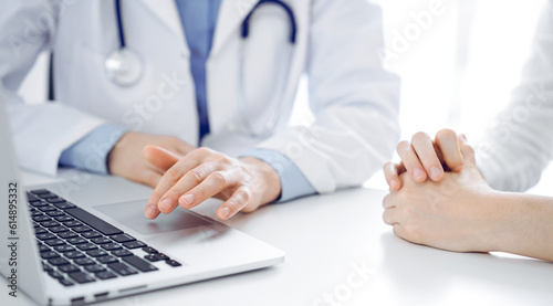 Doctor and patient sitting near each other at the desk in clinic. The focus is on female physician s hands using laptop computer  close up. Medicine concept