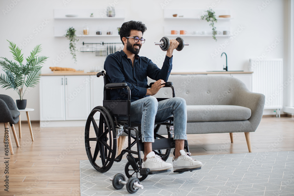 Full length view of young indian male with mobility impairment doing seated exercises with weights in open-plan kitchen. Positive bespectacled adult in jeans gaining health benefits from training. Full length view of young indian male with mobility impairment doing seated exercises with weights in open-plan kitchen. Positive bespectacled adult in jeans gaining health benefits from training.