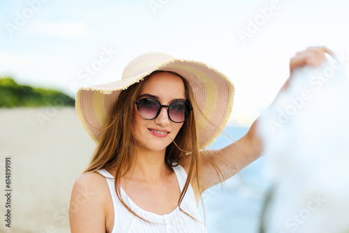 Happy smiling woman in free happiness bliss on ocean beach standing with hat, sunglasses, and white flowers. Portrait of a multicultural female model in white summer dress enjoying nature during