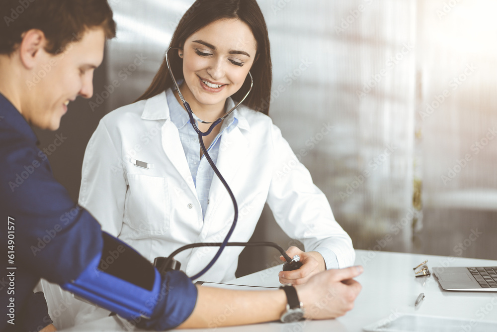 Fototapeta premium Young woman-doctor is checking her patient's blood pressure, while sitting together in clinic. Medicine and healthcare concepts
