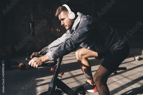 Muscular man in headphones riding exercise bicycle in gym