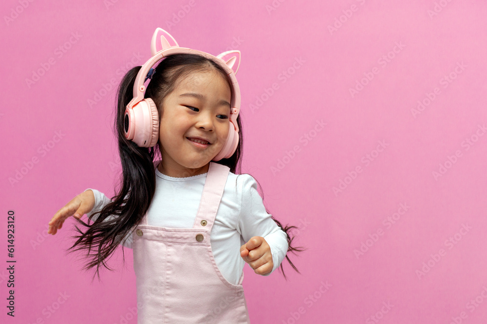 little asian girl in pink children's headphones listens to music and dances on pink isolated background, korean child little asian girl in pink children's headphones listens to music and dances on pink isolated background, korean child