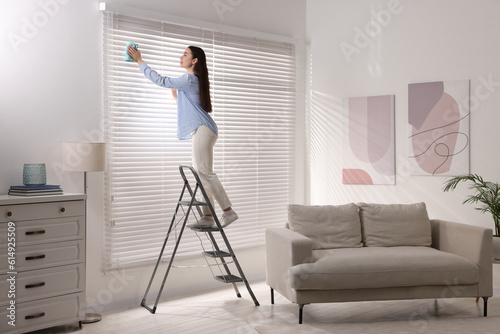 Woman on metal ladder wiping blinds at home