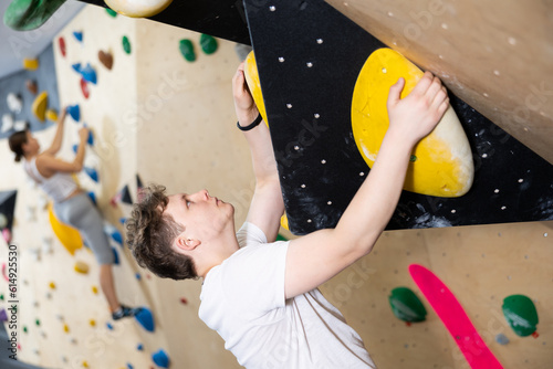 Teenage boy is mastering climbing on training wall in gym, side view. Teen guy holds on tightly to ledges and strives for top of bouldering route