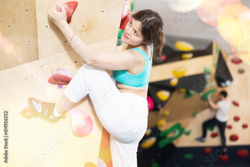 Asian girl is mastering climbing on training wall in gym, side view. Woman holds on tightly to ledges and strives for top of bouldering route