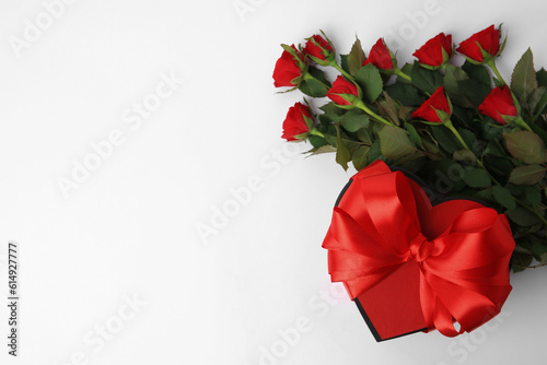 Heart shaped gift box with bow and beautiful red roses on white background, flat lay. Space for text