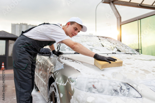 young guy in uniform car wash worker washes car with sponge with foam