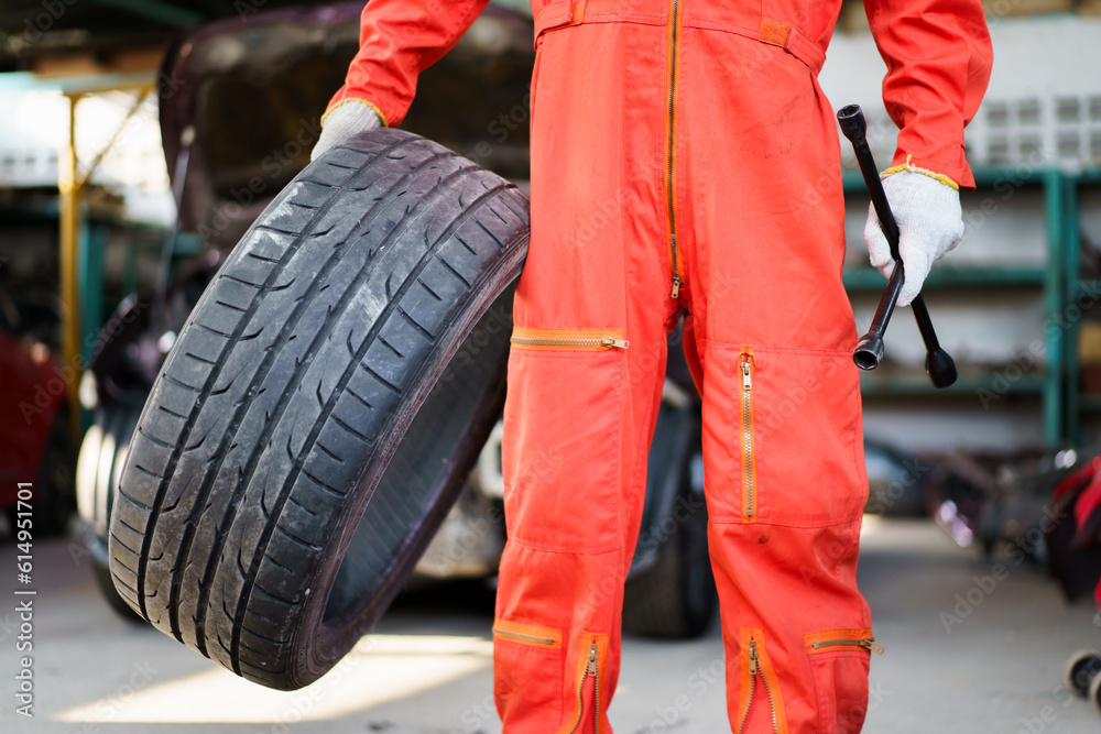 Portrait of professional Asian male vehicle technician or repairman in orange suit holding - carrying a damaged vehicle tyre to be a fixed, a repair man fixing and recap a broken tyre in garage. Portrait of professional Asian male vehicle technician or repairman in orange suit holding - carrying a damaged vehicle tyre to be a fixed, a repair man fixing and recap a broken tyre in garage.