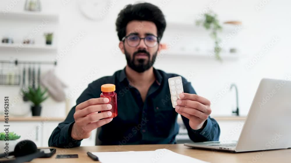 Focus on medicine containers held by arabian young man looking through information on laptop screen in kitchen. Remote employee suffering from stuffy nose during allergy season at work from home. Focus on medicine containers held by arabian young man looking through information on laptop screen in kitchen. Remote employee suffering from stuffy nose during allergy season at work from home.
