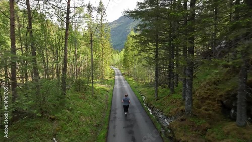 Adult man jogs through forest on paved road. Aerial view following from behind, 60fps photo