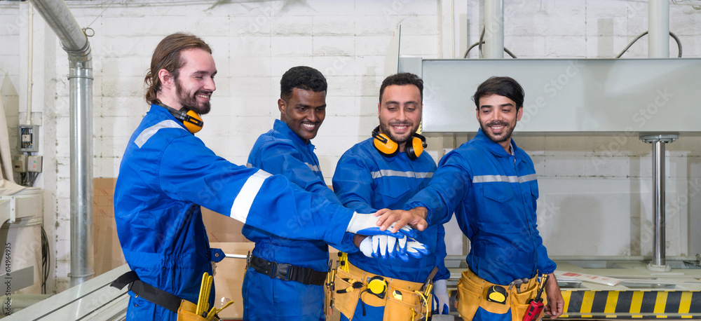 Obraz premium Group of technician in blue mechanic jumpsuit, ear muff and yellow tool belts join hand together in front of polywood cold press machine. Production line atmosphere in furniture factory