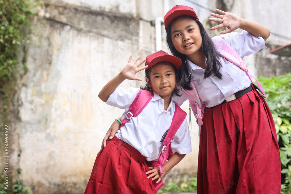 Happy Indonesian schoolgirls of elementary school wearing uniform and backpack waving hand look at camera