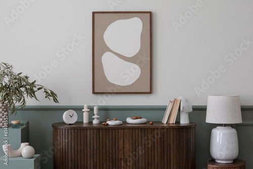 Warm and cozy composition of spring living room interior with mock up poster frame, wooden sideboard, books, plants in flowerpots, stylish lamp and personal accessories. Home decor. Template.