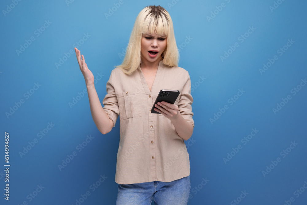 surprised blonde young woman with open mouth looks into smartphone on background with copyspace surprised blonde young woman with open mouth looks into smartphone on background with copyspace