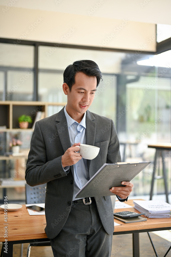 A smart Asian businessman reading document and sipping coffee while standing in the office. A smart Asian businessman reading document and sipping coffee while standing in the office.