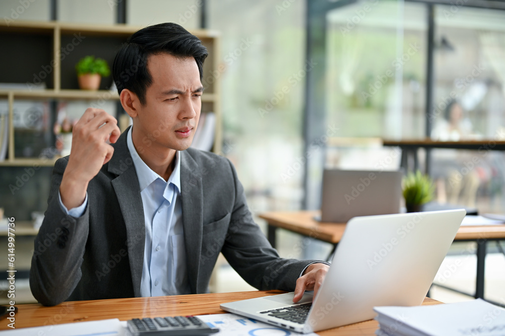 A thoughtful Asian businessman focuses on his tasks on his laptop, working at his desk A thoughtful Asian businessman focuses on his tasks on his laptop, working at his desk
