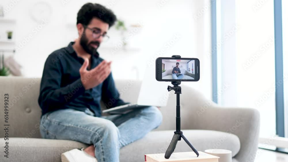 Blurred background of cheerful arabian man sitting on couch with laptop and talking something on smartphone camera. Positive male blogger recording video for his social media. Blurred background of cheerful arabian man sitting on couch with laptop and talking something on smartphone camera. Positive male blogger recording video for his social media.