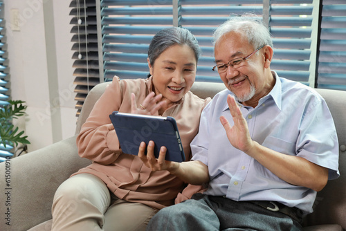 Happy smiling asian senior couple sitting on sofa and using tablet while online video call with friend or relative cousin at home living room. Internet information technology and lifestyle concept.