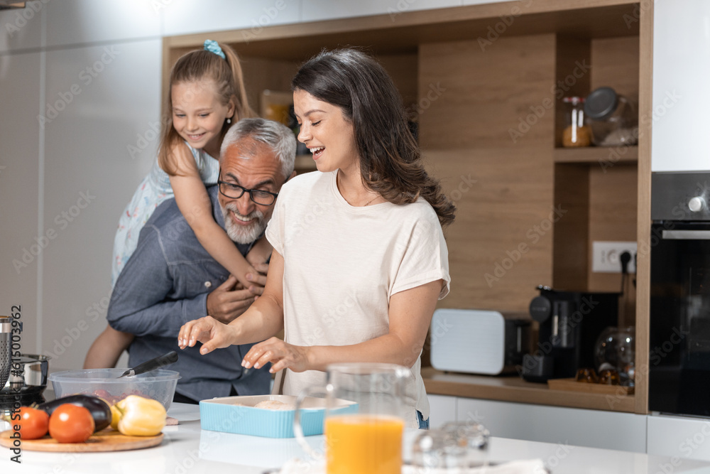Young woman laughing and preparing food as her feather plays with his granddaughter in the kitchen Young woman laughing and preparing food as her feather plays with his granddaughter in the kitchen