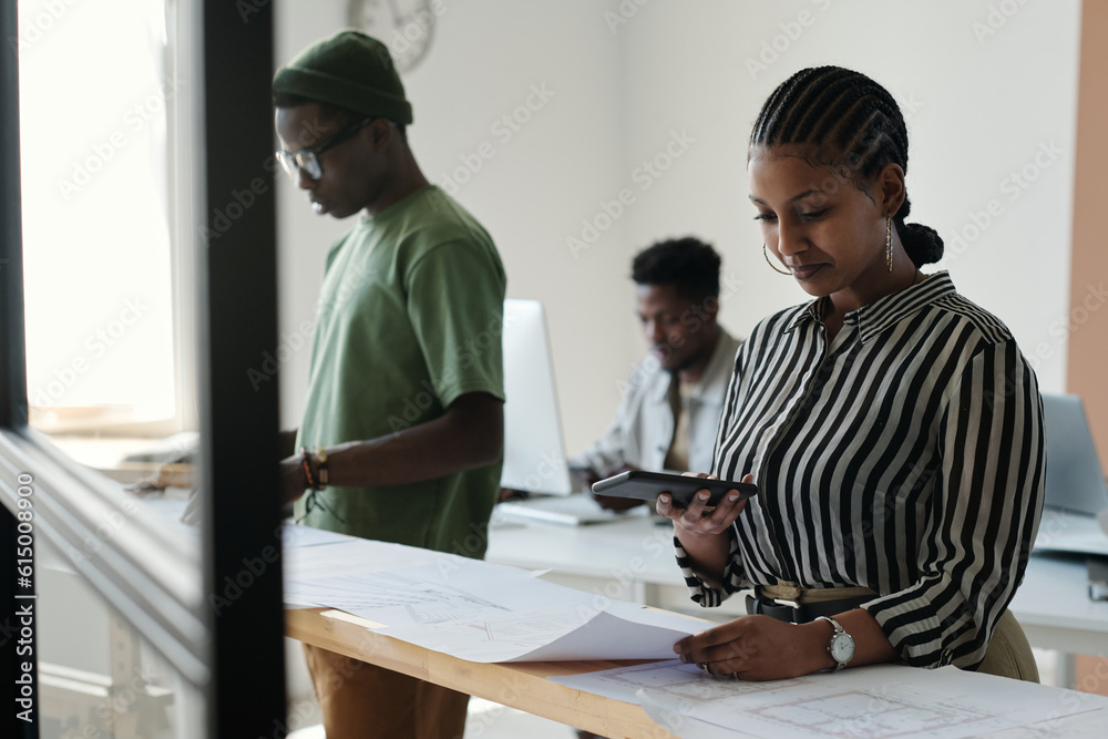 Young businesswoman with smartphone texting or taking photo of blueprint while standing by desk and working with sketches