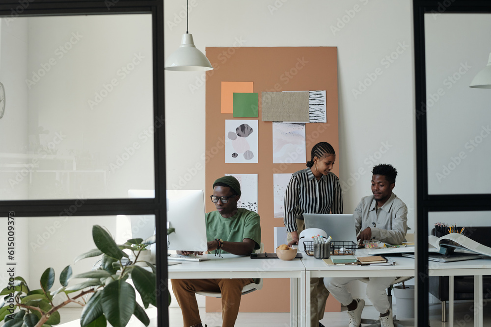 Two young colleagues preparing presentation in front of laptop while sitting next to creative male designer drawing graphic sketches Two young colleagues preparing presentation in front of laptop while sitting next to creative male designer drawing graphic sketches