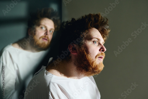 Side view of young insane man in white straitshirt sitting in front of camera against his reflection and looking forwards with anxious expression