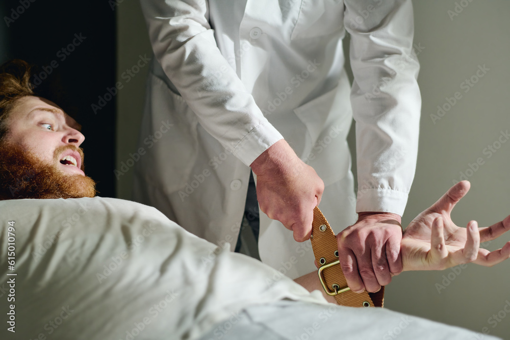 Obraz premium Cropped shot of psychiatrist binding hands of male patient with tight brown belts to calm him down during mental treatment in hospital