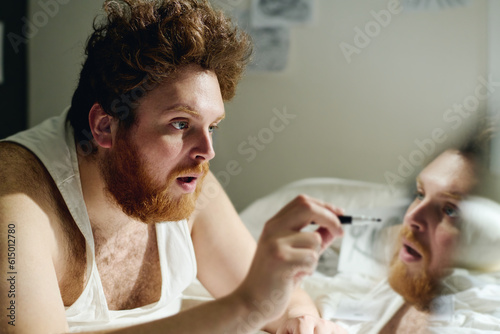 Young fat bearded man with depression or narcissistic mania bending ober bed in front of camera and pointing at reflection of his face photo