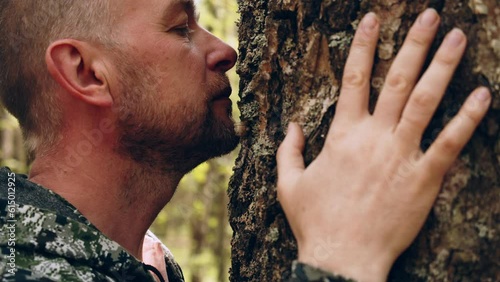 A man walks up to a huge tree, sniffs the bark and hugs the trunk. An adult man enjoys being alone with nature in the forest. The concept of nature conservation, forests, ecology. Human and nature. photo