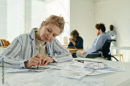 Mature woman with mental disorder drawing something with pencil during course of psychotherapy while sitting by desk in hospital ward