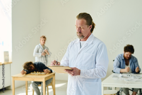 Mature professional pstchiatrist in lab coat making prescriptions for patients in medical document while standing in front of camera