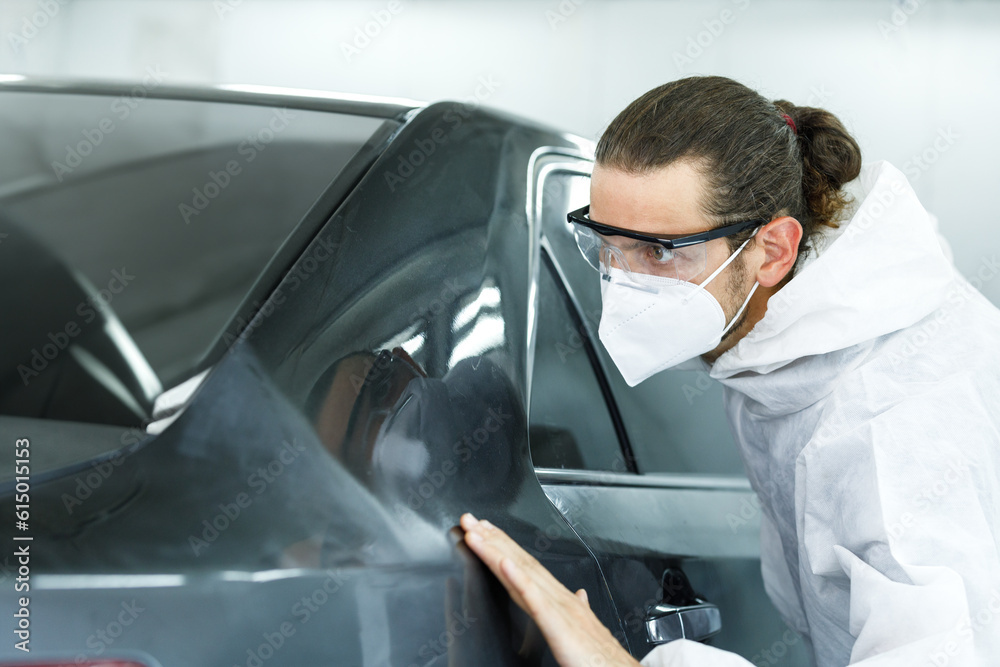 Young professional car painter wearing protective suit standing next to car in car painting room inspecting car body to look for scratches and damage Young professional car painter wearing protective suit standing next to car in car painting room inspecting car body to look for scratches and damage